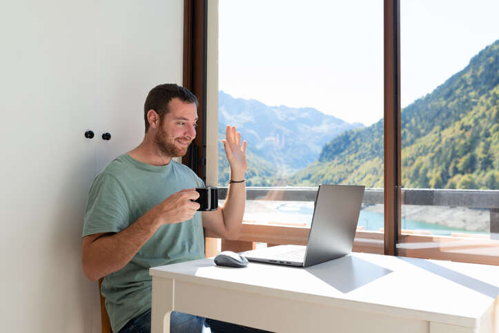 A remote worker takes part in a video conference, with a landscape view in the window behind him.