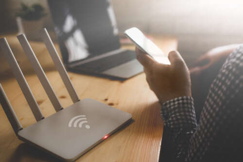 An image of someone holding a mobile phone in front of a laptop and next to an internet router.