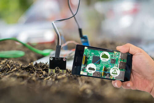 Farmer's hand holding smart phone with agritech icons and messages on screen with soil sensor to manage water, soil quality and monitor weather.