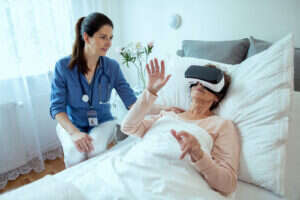 Female Doctor Checking on Elderly Patient Lying in Hospital Bed Doing Therapy Via VR Technology. Friendly Doctor Helping Senior Woman Receive Therapy Using Virtual Reality Headset.