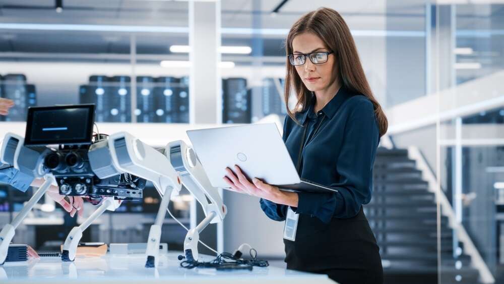 Photo of a woman looking at a laptop next to a robotic device, used to illustrate a story about a new standard on implementing AI in the workplace published by the BSI.
